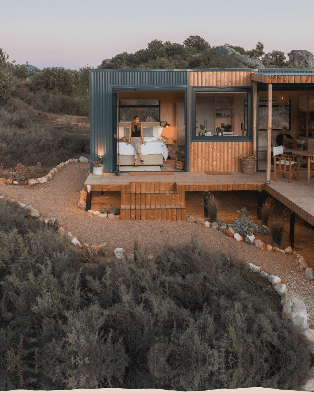 A modern elevated cabin with dark teal metal siding and warm wood paneling, shown at dusk with a person sitting on a bed inside and an outdoor deck in front. The cabin is surrounded by a gravel path, rocks, and dense shrubs in a natural landscape.
